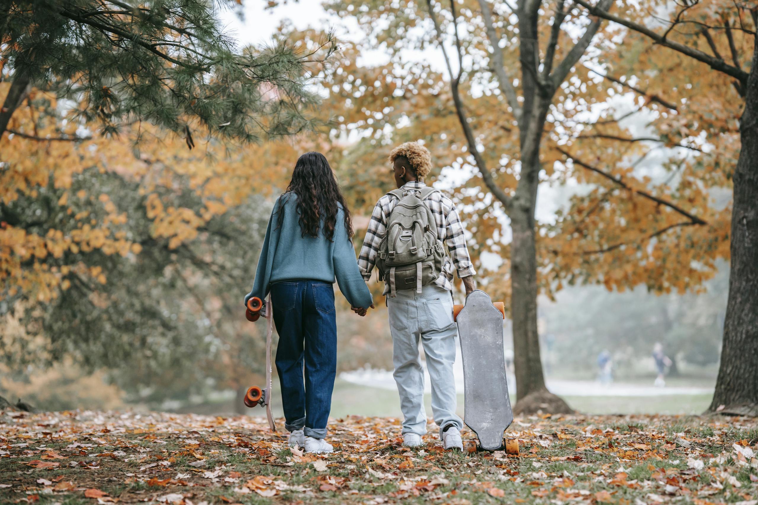 A young couple holding hands and carrying skateboards in a vibrant autumn park setting.