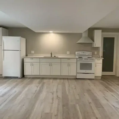 Bright basement kitchenette with light wood‑look floors, greige walls, and a straight run of white shaker cabinets featuring a white fridge, sink, electric range with hood, and a glass‑panel door at the right